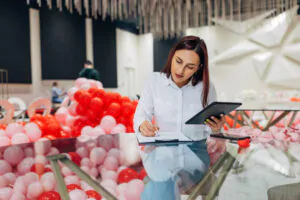 Female event organizer with digital tablet and clipboard, sitting at table in restaurant and writing down new reservations. Organization of parties, ceremonies
