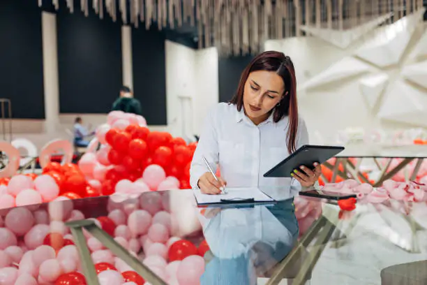 Female event organizer with digital tablet and clipboard, sitting at table in restaurant and writing down new reservations. Organization of parties, ceremonies
